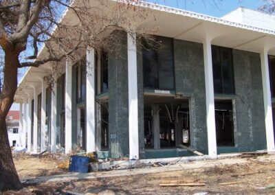The Gulfport Library’s façade shows shattered windows, stripped walls, and debris scattered beneath bare, storm‑damaged trees. The building’s tall white columns remain standing despite extensive destruction.