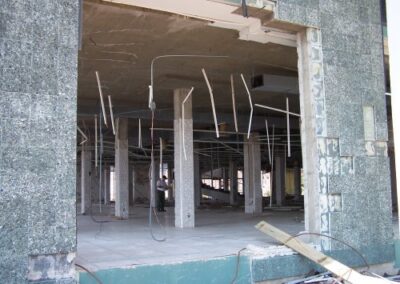 A large opening in the Gulfport Library exterior reveals the stripped interior with hanging ceiling rails, bare columns, and debris covering the floor. Broken wall tiles and structural damage frame the scene.
