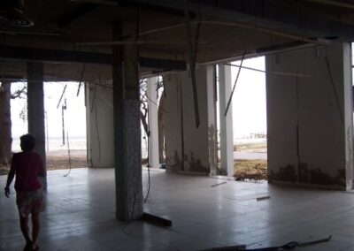 The gutted interior of the Gulfport Library shows bare columns, hanging wires, and mud‑stained walls opening to a bright exterior view. A person stands inside the empty structure surrounded by storm‑damaged flooring.