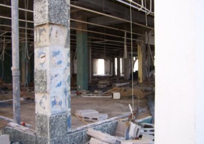 The interior of the Gulfport Library is stripped to concrete floors and columns with scattered cinderblocks, broken framing, and ceiling pieces hanging loose. Light enters through missing walls, revealing extensive structural destruction.