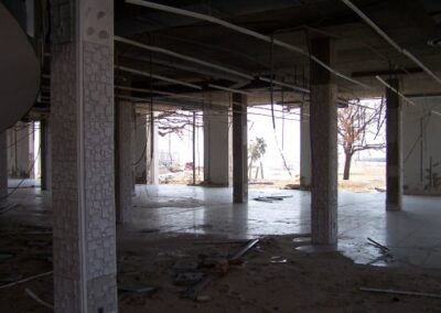 The empty lower level of the Gulfport Library displays tiled floors covered in dirt and debris, with columns and dangling ceiling supports throughout. Open wall sections reveal damaged grounds outside.