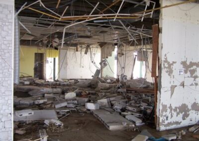 A wide room inside the Gulfport Library shows collapsed ceiling grids, exposed wiring, cinderblocks, and structural debris covering the muddy floor. Walls show peeling paint and severe flood damage.