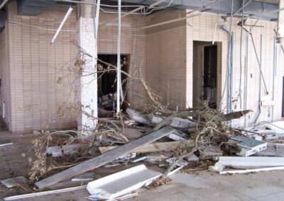 Tree limbs, broken boards, and concrete debris are piled in front of a damaged brick wall inside the Gulfport Library. Ceiling pieces and metal supports hang down from the exposed structure above.