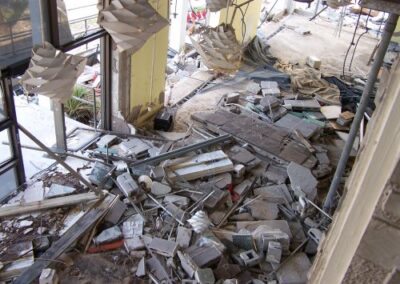 A collapsed interior area of the Gulfport Library is filled with shattered ceiling panels, broken wall sections, and piles of concrete and debris scattered across the floor. Large hanging light fixtures dangle from the damaged ceiling above the destruction.