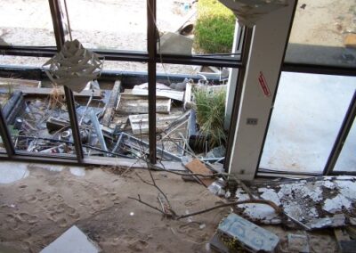 An upper‑level view through large windows shows rubble, broken boards, and storm debris piled against the exterior of the Gulfport Library. Inside, sand and damaged materials cover the floor near the glass wall.