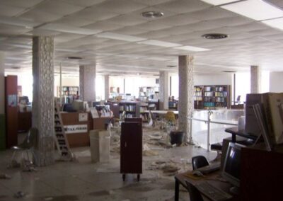 The upper floor of the Gulfport Library shows scattered debris, damaged ceiling tiles, and disorganized furniture, with bookshelves still standing in the background. Mud and paper fragments cover the tiled floor.
