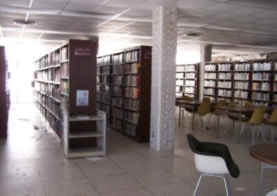 Rows of tall bookshelves remain standing inside the Gulfport Library, with tables and chairs nearby, while sections of ceiling tiles are missing or damaged. The tiled floor shows scattered dirt and storm residue.