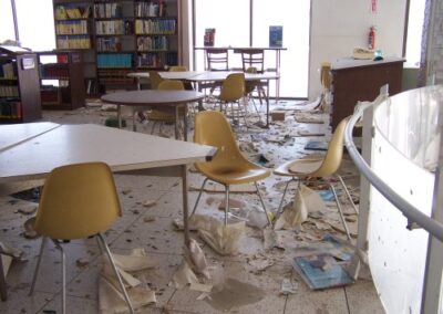 A reading area inside the Gulfport Library shows scattered chairs, tables, books, and papers strewn across the floor. Water damage and debris are visible beneath bookshelves near large windows.