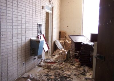 A hallway inside the Gulfport Library is filled with broken furniture, soaked papers, and debris piled along the walls and floor. Light enters through a tall window at the end of the corridor, revealing extensive interior damage.