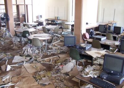 Rows of tables, chairs, and damaged desktop computers sit among piles of wet books, ceiling tiles, and debris covering the library floor. The destruction extends throughout the open reading area.