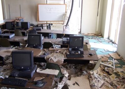 A computer lab inside the Gulfport Library is filled with mud‑soaked papers, collapsed ceiling material, and overturned furniture surrounding rows of damaged desktop computers. Sunlight filters through missing sections of wall at the back of the room.