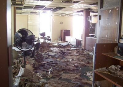 A large room in the Gulfport Library is strewn with soaked books, fallen ceiling tiles, collapsed furniture, and debris covering nearly the entire floor. A large fan sits among the destruction beneath a partially collapsed ceiling grid.