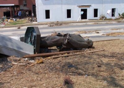 A toppled bronze statue lies on the ground beside its fallen pedestal, surrounded by storm debris on a damaged lawn near the Gulfport Library. Broken boards, scattered materials, and heavily damaged buildings appear in the background.