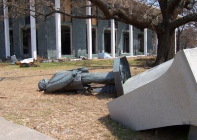 The fallen statue rests on its back near the Gulfport Library, with its heavy stone pedestal overturned beside it. The damaged library building and a large oak tree stand behind the scattered hurricane debris.