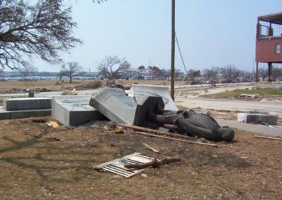 A toppled bronze statue lies beside its fallen pedestal on a debris‑littered lawn near the Gulfport Library, with damaged buildings and scattered materials in the background. Broken boards and storm wreckage surround the figure.