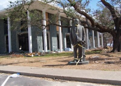 The same statue has been lifted upright and secured with straps in front of the storm‑damaged Gulfport Library. Fallen branches, debris, and missing windows highlight the surrounding destruction.