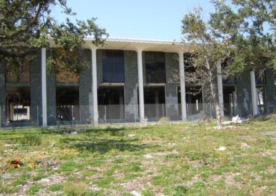 A wide view of the Gulfport Library shows its tall façade with missing windows, boarded sections, and a debris‑strewn lawn. Trees around the building are stripped and damaged from hurricane winds.