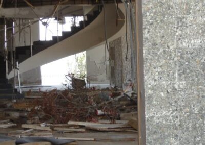 The interior of the Gulfport Library shows a curved staircase rising behind piles of storm debris, broken ceiling materials, and exposed wiring. Light enters from the far side of the room, illuminating the heavily damaged space.