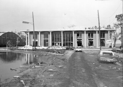 A black‑and‑white exterior view of a modern library building with large windows, surrounded by debris, mud, and standing water after severe storm damage. Several cars are parked near the entrance with visible destruction in the surrounding area.