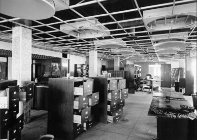 A library interior showing rows of open cabinets and scattered files beneath a partially dismantled ceiling grid. Various pieces of furniture and shelving are pushed into uneven positions as the room undergoes cleanup.