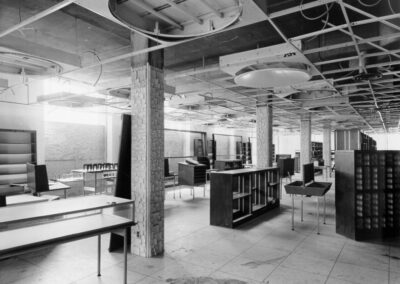 A black‑and‑white photo of a library interior with ceiling panels removed, exposed wiring, scattered shelving, and furniture displaced across the room. The space appears mid‑cleanup or repair following structural damage.