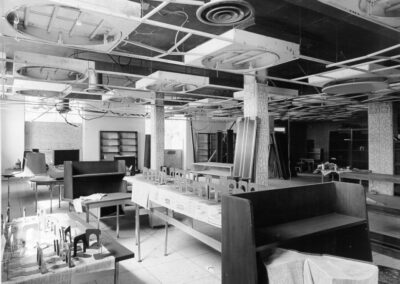 A damaged library interior with ceiling tiles missing, debris on tables and floors, and furniture pushed into disorganized arrangements. Light filters through uncovered windows, illuminating the cluttered room.