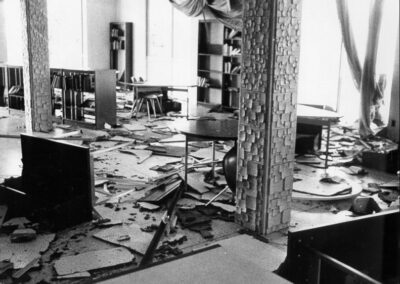 A black‑and‑white photo of a heavily damaged library interior with collapsed ceiling pieces, scattered debris, broken furniture, and books displaced across the floor. Support columns and shelving remain standing amid the destruction.
