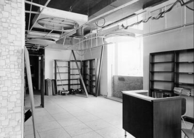 A damaged library room with empty bookshelves, exposed ceiling framing, and leaning ladders and panels stacked near the back wall. Light enters through a large window, highlighting the disarray of the space.