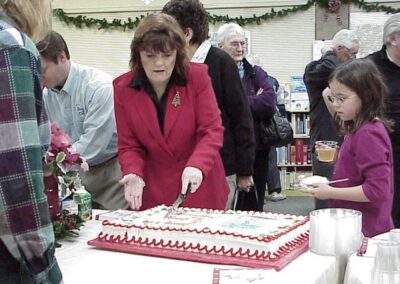A woman cuts a large decorated sheet cake during a community gathering inside a library. People stand in line behind the table as refreshments are served.