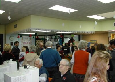 A library filled with holiday decorations hosts a gathering where people stand and converse near bookshelves and service desks. Snowflake cutouts and festive garlands hang from the ceiling.