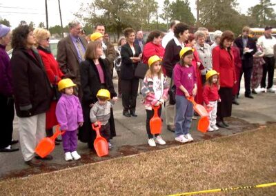 Children wearing yellow hard hats and holding plastic toy shovels stand with a group of adults at an outdoor groundbreaking event. The attendees form a line facing the speaker’s area near a grassy patch.