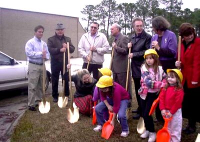 A group of adults and children, some wearing yellow hard hats, pose together outdoors holding ceremonial and toy shovels. The setting appears to be a grassy area beside a parking lot.