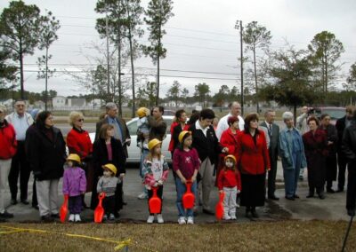 A crowd of families and community members, including children holding toy shovels, stands outdoors on a paved area near a grassy lawn during a groundbreaking ceremony. Trees and utility lines are visible in the background.