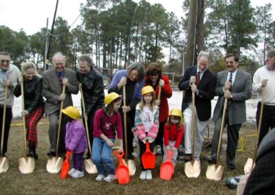 Adults and children wearing yellow hard hats gather outdoors holding gold ceremonial shovels and bright plastic toy shovels. The group stands on grass near a parking area during a groundbreaking event.