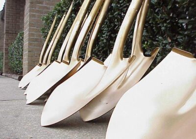 A row of gold ceremonial shovels leans against a low hedge beside a building wall. The shovels are arranged closely in a diagonal line.