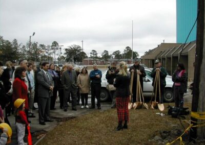 A group of people gathers outdoors as a woman with a microphone addresses the crowd near a line of ceremonial gold shovels. Attendees stand on a paved area beside a lawn under an overcast sky.