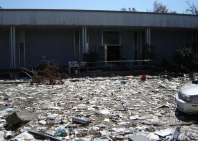 The area in front of the Pass Christian Library is covered with a thick layer of papers, books, and debris washed out by the storm surge. The entrance sits behind ruined vegetation and broken materials.