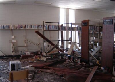The interior of the Pass Christian Library is filled with collapsed bookshelves, scattered books, broken furniture, and mud‑covered flooring. Overhead fluorescent lights and ceiling tiles hang askew.