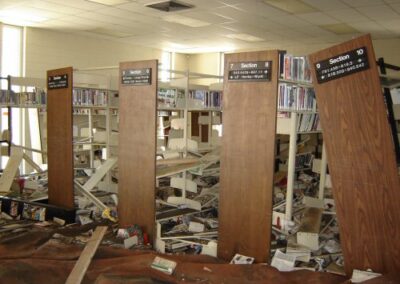 Rows of labeled shelving end panels stand amid collapsed bookcases, scattered books, and storm debris covering the library floor. Ceiling damage and warped carpet indicate severe flooding.