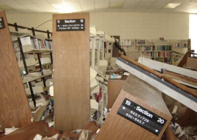 Collapsed bookshelves, broken furniture, and scattered books fill the interior of the Pass Christian Library. Upright section end panels remain standing among the extensive storm debris.