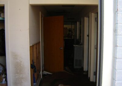 A narrow hallway inside the Pass Christian Library shows an open door leading into a dark, damaged room, with mud‑stained floors and partially removed wall sections. Debris and fallen ceiling material line the corridor.