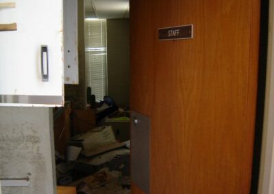 A door labeled “STAFF” stands open, revealing a cluttered, debris‑filled room with damaged furniture and scattered materials. The surrounding walls and floor show heavy water staining.