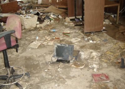 A muddy library floor is scattered with debris, broken electronics, and damaged books, with pieces of furniture leaning at odd angles. A torn office chair sits nearby among the destruction.