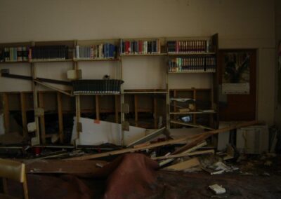 Collapsed shelves, torn carpet, and scattered books cover the interior of the Pass Christian Library. The back wall’s shelves remain partially intact above the storm‑damaged floor.