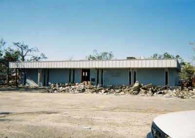 A wide view of the Pass Christian Library shows a long, low building surrounded by piles of rubble and debris across the front lot. Trees are stripped bare, and the structure shows significant storm damage.