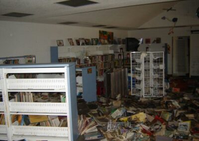 Rows of white wire racks and bookshelves stand among piles of soaked books and debris inside the Pass Christian Library. The ceiling panels above are stained or missing from storm damage.
