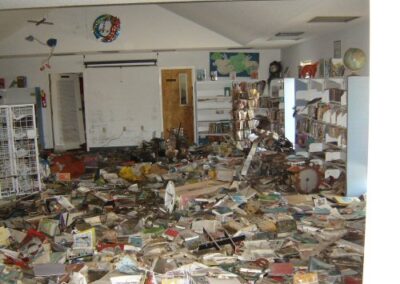 The children’s area of the Pass Christian Library is filled with piles of ruined books, collapsed shelves, and storm debris scattered across the floor. Educational posters and decorations remain hanging above the destruction.