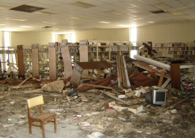 The main library floor of the Pass Christian Library is covered with collapsed shelves, broken furniture, and scattered books. A lone wooden chair sits amid the mud‑stained debris.