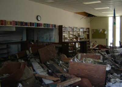 A heavily damaged interior of the Pass Christian Library shows collapsed shelves, broken furniture, and scattered debris across the floor. Wall shelves remain partially intact above piles of storm‑soaked materials.