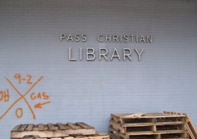 The exterior wall of the Pass Christian Library shows its signage above storm debris and spray‑painted search‑and‑rescue markings. Stacked wooden pallets sit in front of the damaged brick wall.